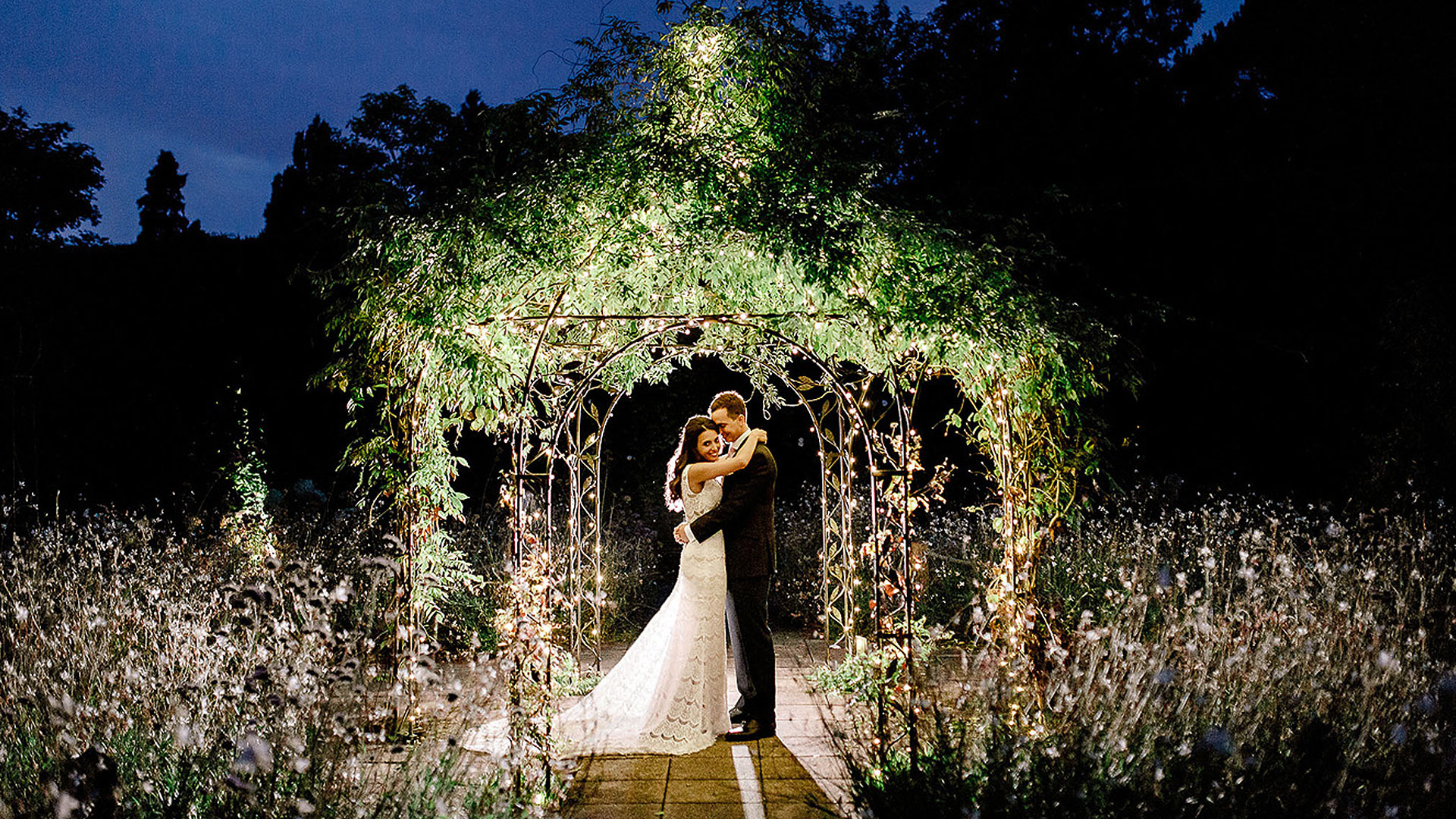A happy couple embrace as they stand under the romantic wrought-iron pavilion at night - wedding photo ideas