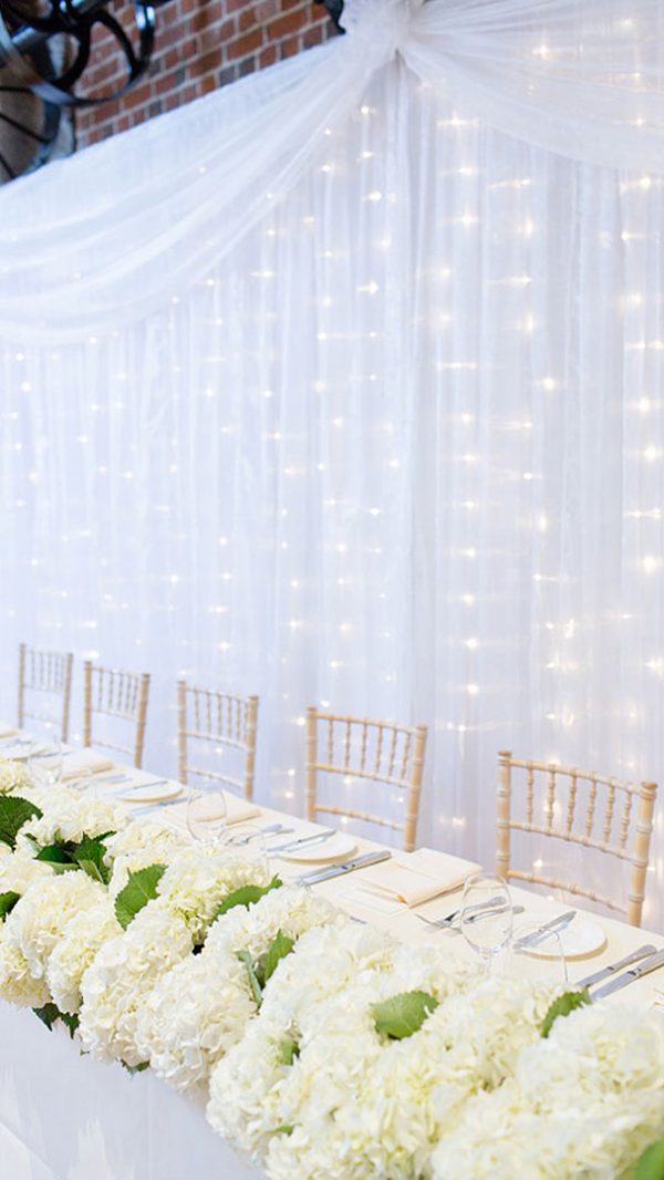 A white curtain with fairy lights hangs behind the top table inside Essex wedding barn