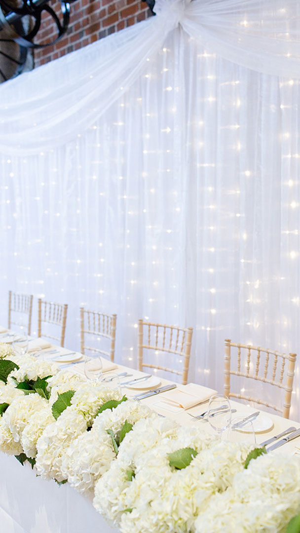 A white curtain with fairy lights hangs behind the top table inside Essex wedding barn