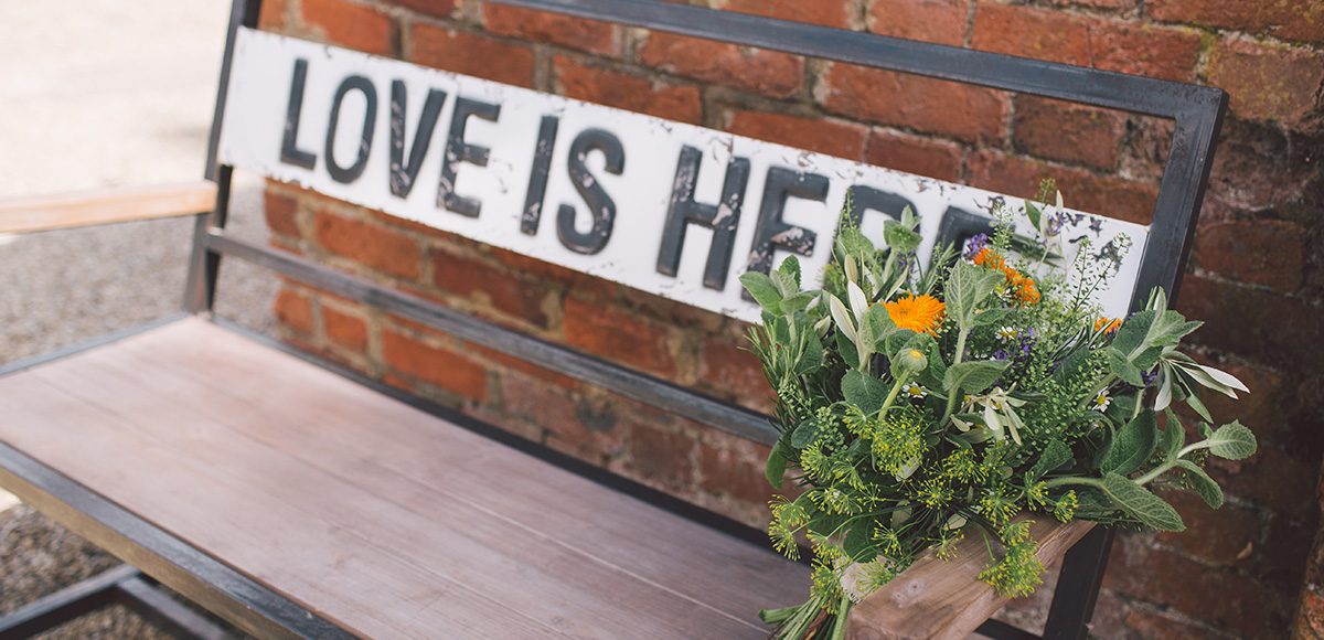 A close up of a wedding bouquet placed on a bench with a sign that reads ‘Love is here’
