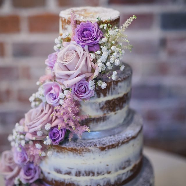 A four-tiered semi-naked wedding cake is decorated with pink and purple roses