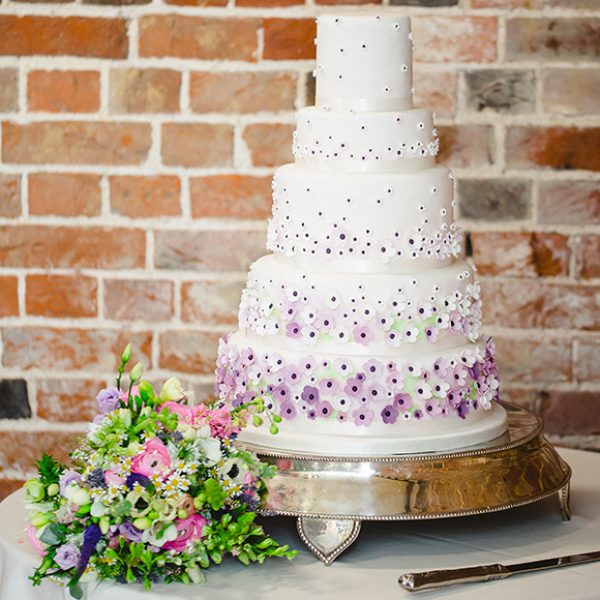 A five-tiered traditional wedding cake is decorated with small purple and white icing flowers