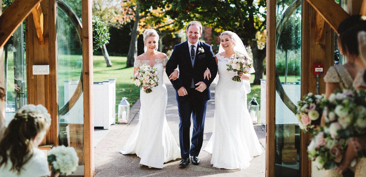 The brides walk with one of their fathers down the wedding aisle towards the Orangery at Gaynes Park