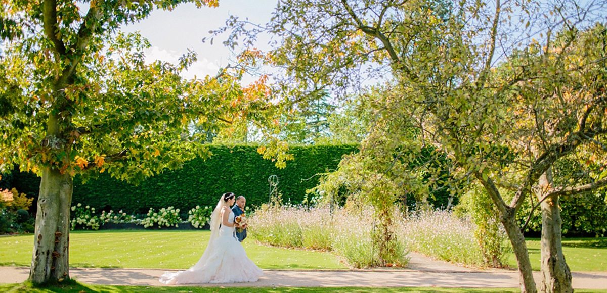 The bride and her father walk down the wedding aisle in the walled gardens at Gaynes Park in Essex